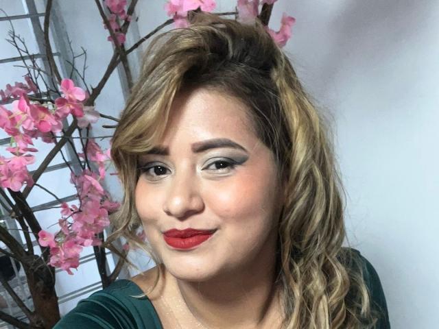 CoolSweets has wavy blonde hair, red lipstick, and green eyes with smoky eyeliner. She's smiling with pink cherry blossoms in the background. Her tits are not visible.