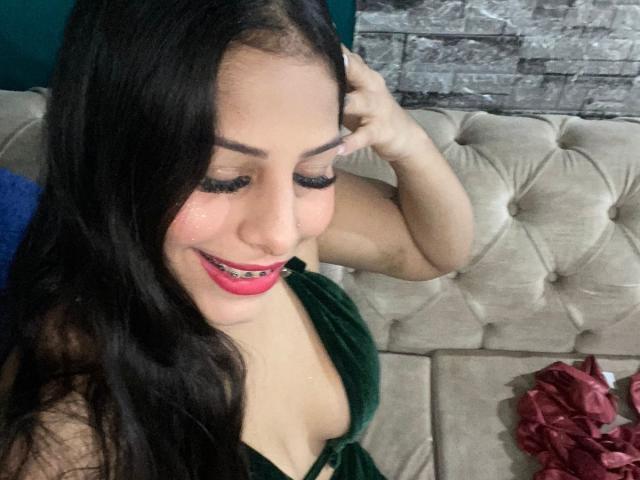 CoolSweets with long black hair, pink lipstick, and green dress, showing off her tits, smiling with eyes closed, hand in hair, sitting on beige tufted couch.