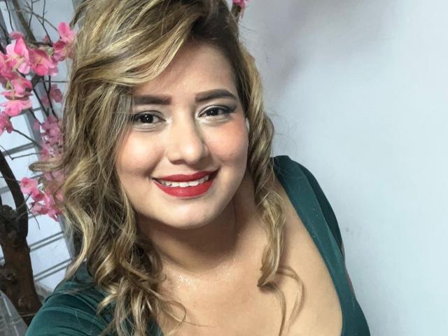 CoolSweets with wavy blonde hair, red lipstick, green dress, smiling, showing her tits, pink flowers in background, white wall, soft light, cute, sexy.