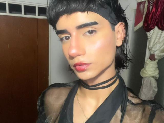 IvyMiller has dark eyebrows, short black hair with a headband, red lips, shiny skin, black choker, and sheer black top, showing a slim, androgynous face.
