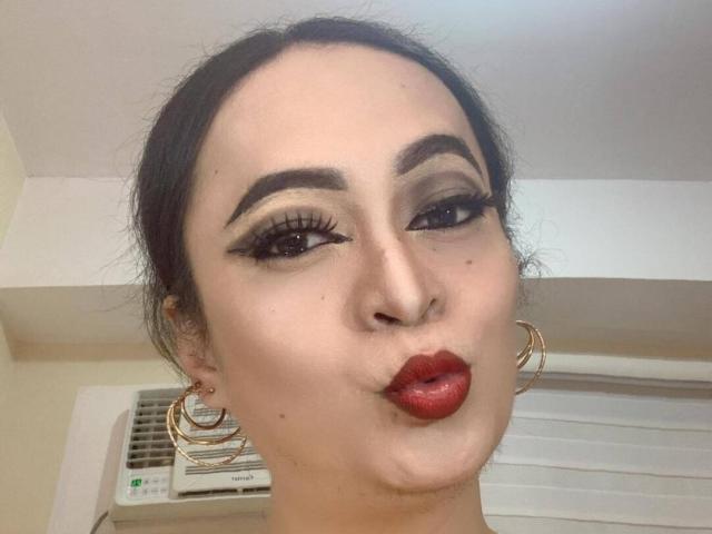SquirtCumAlots has dark hair, thick eyebrows, red lipstick, and large gold hoop earrings. She's making a pouty, puckered-up face with long eyelashes and a slight smile.