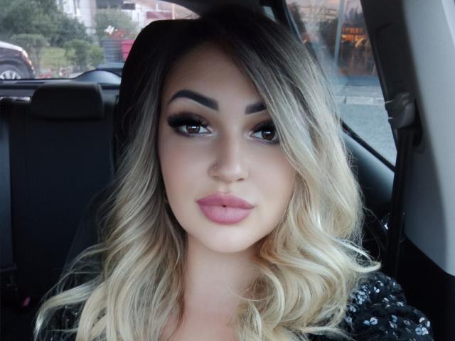 CherryVelvetRed has blonde, wavy hair, full pink lips, and thick, dark eyebrows. She's wearing black eyeliner and mascara, looking straight into the camera with a neutral expression.