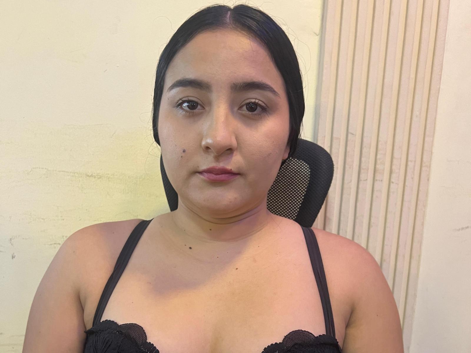 ValeriaTurner with black hair, brown eyes, and a black lace top, sitting in a chair, showing a neutral expression, with a small mole on her left cheek.