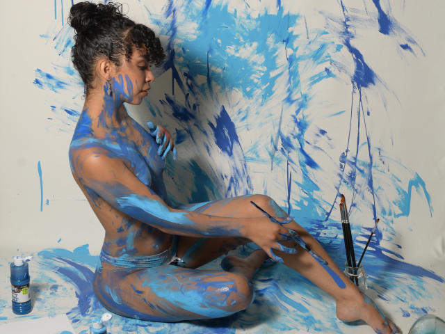JadeMiranda, naked with blue paint covering her tits, ass, and legs, sits on the floor, painting brush in hand, with more paint splattered around her.