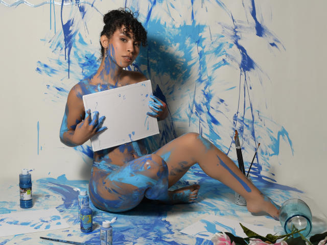 JadeMiranda with blue paint splattered on her brown skin, curly hair, and nude body, holding a white sheet, sitting on the floor, surrounded by paint bottles and brushes.