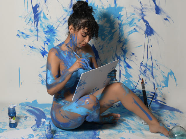 JadeMiranda, naked, covered in blue paint, sits with a tablet, painting, holding a brush, curly hair in a bun, paint splashes on her tits, ass, and legs, white background.