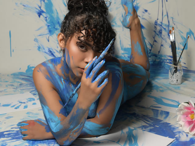 JadeMiranda, a curly-haired woman with tan skin, lies on paint-splattered paper, her blue-painted tits, ass, and hand, covering her mouth with a paintbrush, surrounded by paintbrushes.