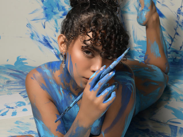 JadeMiranda, curly-haired, tan-skinned, lying on blue-painted surface, holding paintbrush, face and body covered in blue paint, looking at camera, legs raised, paint on feet, earrings visible.