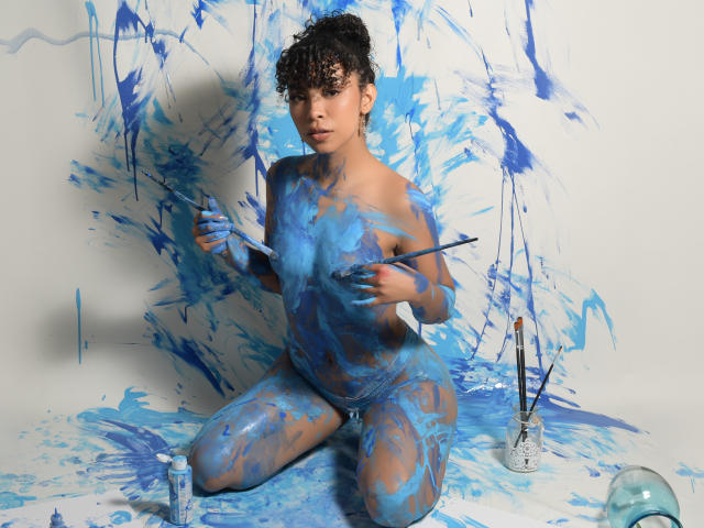 JadeMiranda, nude with blue paint splattered on her body, kneels holding paintbrushes, curly hair, paint bottle and brushes on white floor, looking at camera, paint stains everywhere.
