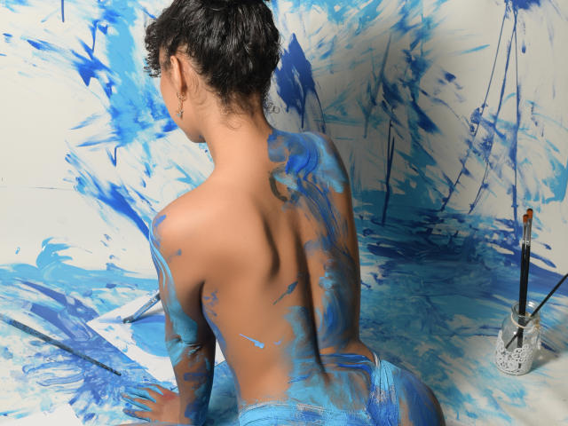 JadeMiranda is topless with blue paint on her back, butt, and arms, sitting with her back to the camera, curly black hair, paintbrushes on the right.