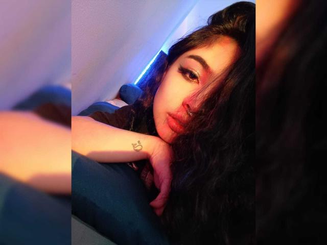 MeghanRider with long black hair, pouty lips, and a small heart tattoo on her right wrist, lying on a blue surface, looking at the camera with a sultry expression.