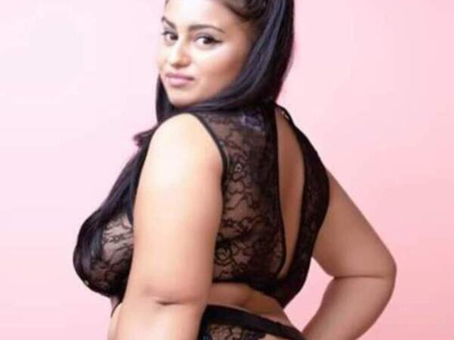 RubyFiera with long black hair, wearing a sheer black lace top, showing her big boobs and curvy ass, looking over her shoulder with a slight smile.