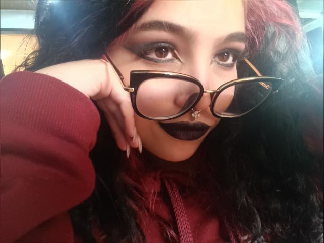 MeghanRider with dark purple lipstick, black curly hair, red highlights, tortoiseshell glasses, nose ring, and red sweater, touching her face with manicured nails, looking at camera.