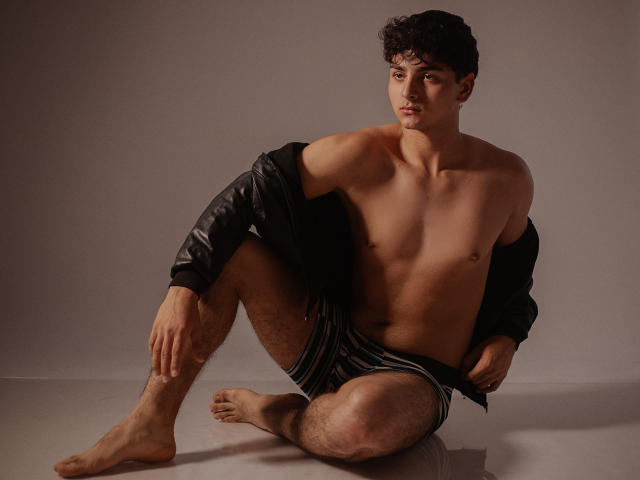 JakeSummer, senza camicia con la giacca di pelle nera appesa, indossa pantaloncini a righe, seduto a terra con le gambe incrociate, capelli ricci neri, muscoloso, pelle abbronzata, guarda la fotocamera, piedi nudi, leggermente attraente..