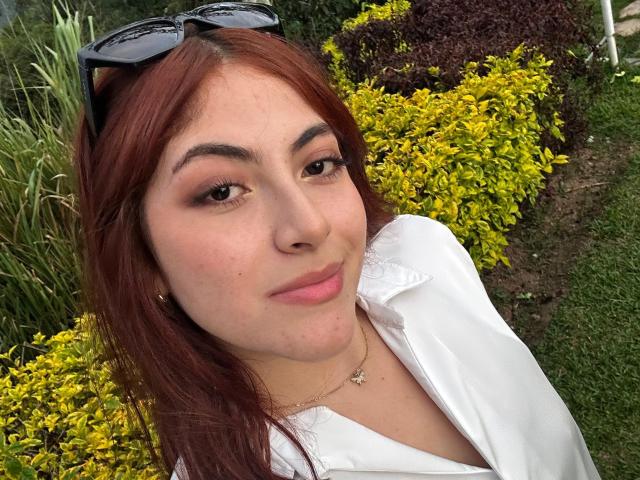 SpencerLe with red hair, white shirt, subtle makeup, small gold necklace, eyeglasses on head, smiling, looking at camera, green bush, yellow foliage in background.