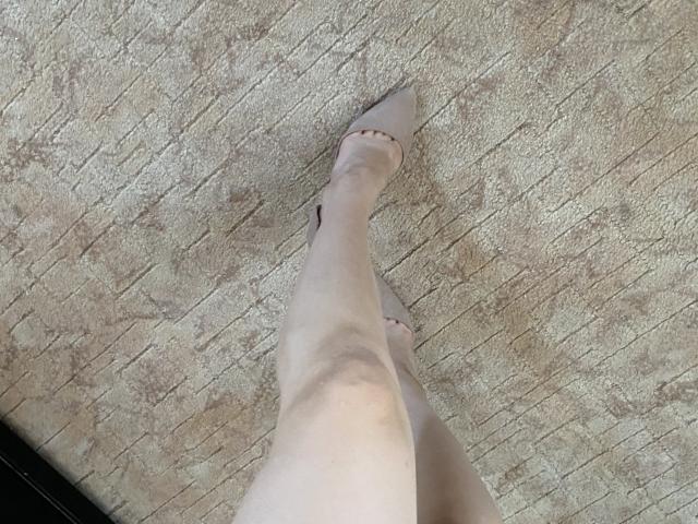 Olieri's been draagt naadloze, transparante kousen en puntige hakken, en staat op een beige tapijt. De enkel is zichtbaar, licht gebogen, en de voet wijst omhoog, waarbij de punt van de hak zichtbaar is..