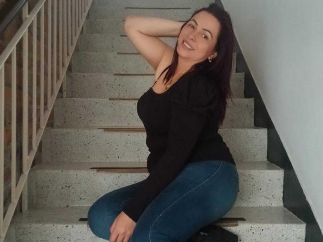 PamelaSmiths has dark brown hair, a black long-sleeve shirt, and blue jeans, kneeling on a speckled stair, smiling with one arm behind her head.