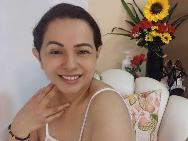 PamelaSmiths with dark hair, smiling, wearing a white tank top, her hand on her neck, sitting on a white couch with floral pillow, yellow sunflower, red flowers in background.