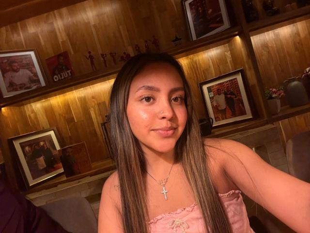 Courttneyy with long brown hair, pink strapless top, cross necklace, slight smile, glowing skin, wooden wall shelf, framed photos, warm lighting, cute, attractive, tits not visible.