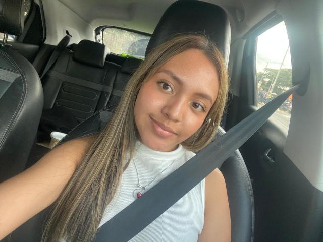Courttneyy with long brown hair, brown eyes, and a small smile, wearing a white sleeveless top, seatbelt across chest, sitting in a car with black leather seats.