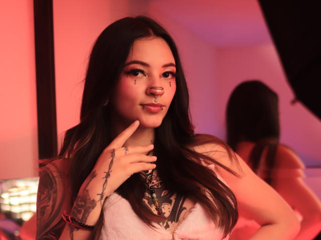 KittsRavee with long dark hair, nose ring, and tattoos, wearing a pink top, touching her chin, smiling, red and pink lighting, mirror reflection in the background.