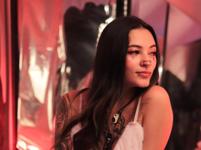 KittsRavee, com cabelo preto longo, tatuagens, piercing no nariz e delineador, sorrindo para a câmera, vestindo uma blusa branca e fofa, com uma luz vermelha ao fundo, parecendo sexy e confiante..