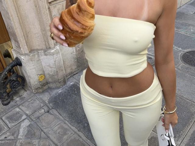 Arabia with tan skin, wearing a yellow tube top and matching pants, holds a croissant in her right hand, showing her bare tits and a flat, toned stomach.