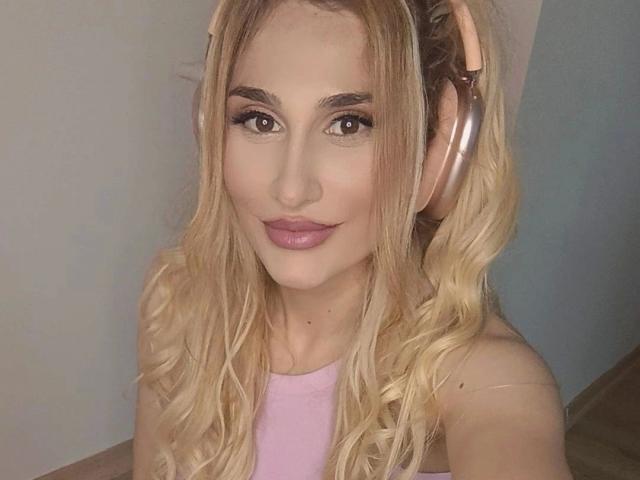 LaurinaxXx, com cabelos loiros ondulados, batom rosa e fones de ouvido cor de rosa, sorrindo para a câmera. Ela está vestindo uma blusa rosa, com os seios quase imperceptíveis..
