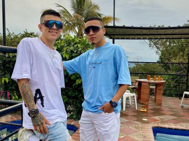 Two dudes with sunglasses, AlejoAlex on left in white tee and ripped jeans, AlejoAlex on right in blue tee and white pants, standing by a pool, hands touching.