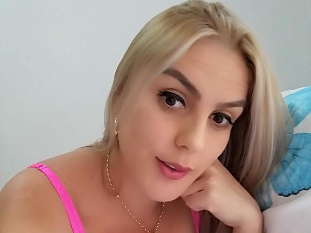 SkarletBlond with blonde hair, pink lipstick, and pink bra, looking at the camera with a slight smile, gold necklace, and hand resting on her cheek.