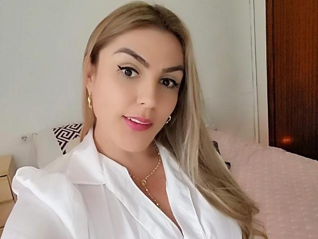 SkarletBlond has blonde hair, wearing a white shirt, gold earrings, and a gold necklace, with pink lipstick and eyeliner, smiling at the camera.