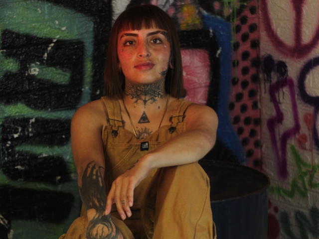 Aisha69Hot with brown bob, tattoos, and nose ring, wearing tan overalls, sitting with arm on knee, red light on face, looking at camera, in graffiti-filled room.