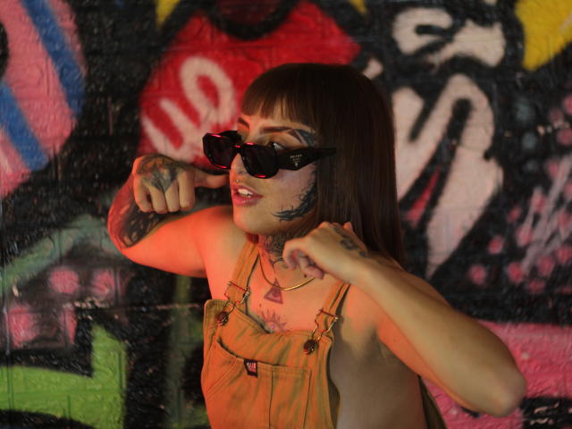 Aisha69Hot with tattoos, wearing black sunglasses, orange overalls, and red lipstick, adjusting her glasses with both hands, showing off her tits and tattoos.