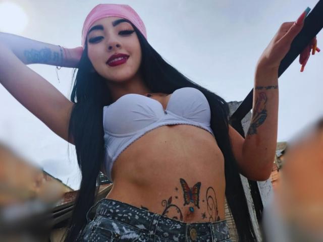 Viiolett with long black hair, pink beanie, white bra showing her boobs, tattoos on her side and arm, and painted red lips, wearing camo pants, looking down at the camera.