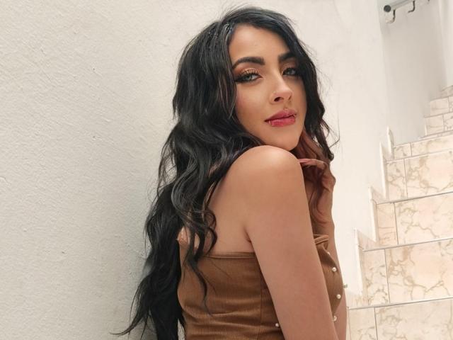 Viiolett with long black wavy hair, brown strapless top, red lipstick, and brown gloves, looking over her shoulder, showing her bare shoulders and upper back.