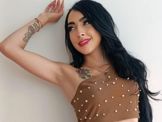 Viiolett with long black hair, red lipstick, and brown, polka-dot crop top, showing tattooed arm and smiling at camera. Her tits are small, her ass is not visible.