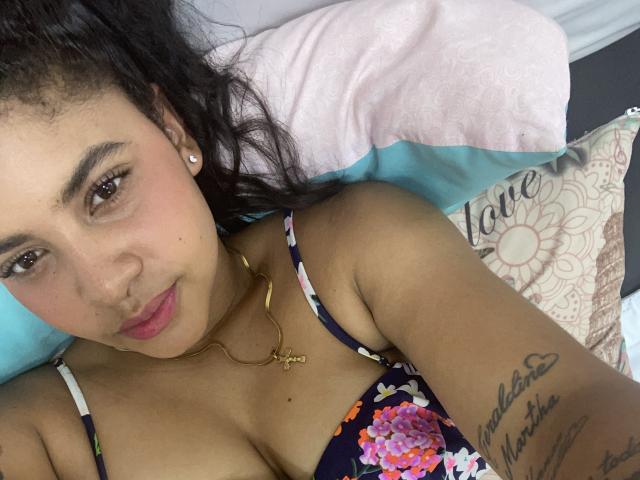 YaraMiler with dark curly hair, pink lips, and brown eyes, wearing a floral dress, gold necklace, and earrings, showing a tattoo on her right arm, lying on a pillow.