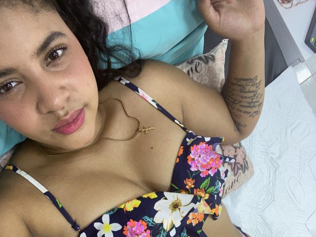 YaraMiler with dark curly hair, brown eyes, and pink lipstick, wearing a floral bra, has a tattoo on her right arm, and a gold necklace, lying in bed.