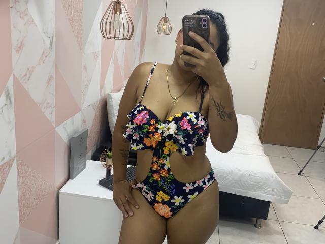 YaraMiler with dark skin, wearing a floral bra and panty set, showing her big boobs and curvy ass, taking a mirror selfie, tattoo on her right arm, holding a phone.