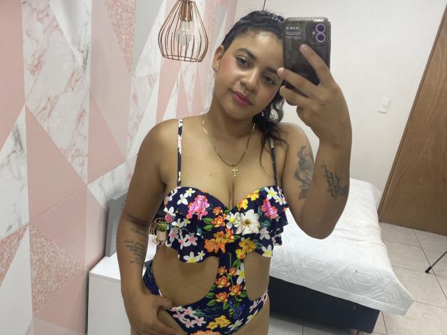 YaraMiler with dark skin and curly hair, wearing a floral bra and panty set, showing her tits and tattoos, taking a selfie in a room with pink geometric walls.