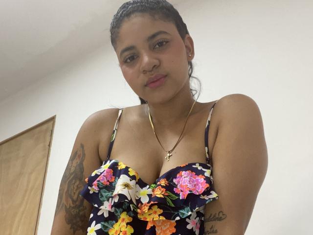 YaraMiler with dark braids, wearing a floral dress with visible cleavage, gold necklace, and tattoos on her right arm, smiling slightly at the camera.