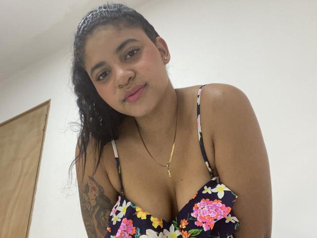 YaraMiler with dark curly hair, brown eyes, nose ring, wearing a floral black bra, showing her tits, tattoo on right arm, gold necklace, smiling softly, in a white room.