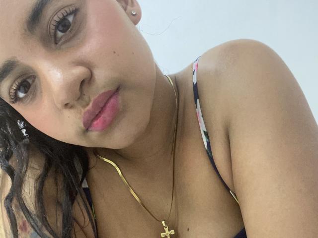 YaraMiler has brown eyes, pink lips, and a gold necklace with a cross. She's wearing a patterned top, showing her bare shoulders and dark, wavy hair.