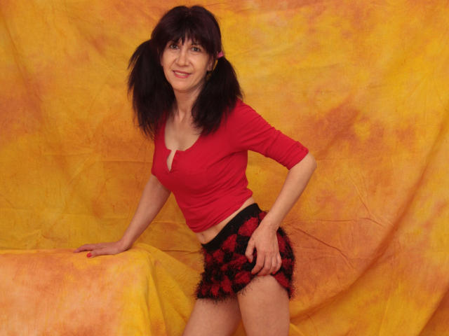 IWannaLoveYou con cabello negro y rizado, una blusa roja y una falda a cuadros negra y roja, sonriendo, apoyada en una tela amarilla, mostrando su trasero en la foto..