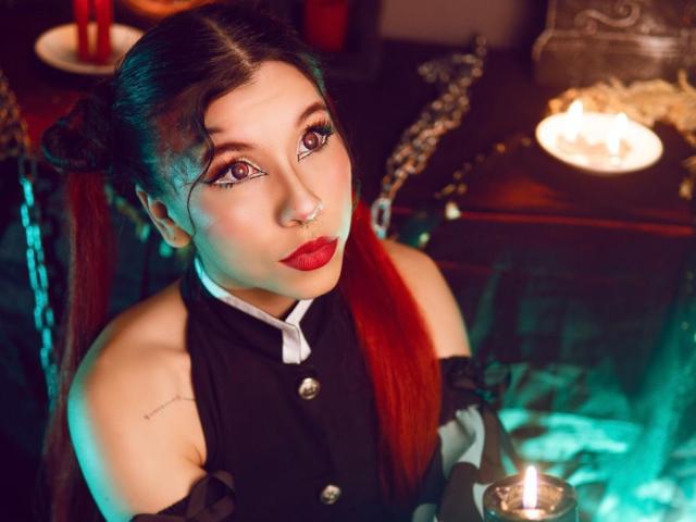 SofiBrandford has red lipstick, black dress with buttons, and a nose ring, holding a candle, with red and black hair in pigtails, and a tattoo on her left shoulder.
