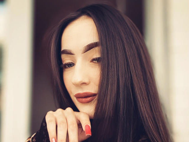 TuttiLily, con i suoi lunghi capelli castani, un occhiolino e il rossetto rosso, ha la mano vicino al viso, con le unghie dipinte di rosso, appare sexy e sicura di sé..