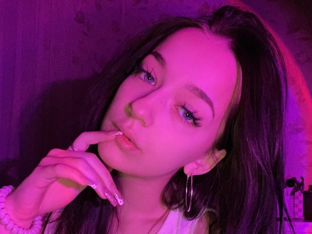 TuttiLily con rostro de tonos rosados, labios carnosos, dedo en la boca, cabello oscuro, ojos azules, aros grandes, coletero blanco, uñas largas, iluminación púrpura, linda, sexy..