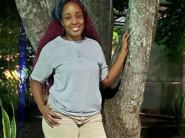 ChelseyeBonny with dark skin, red curly hair, wearing a gray shirt and beige pants, standing against a tree, smiling, hand on hip, other hand on tree.