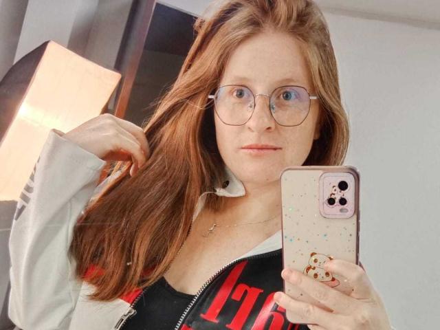 EnzumGinger with red hair, glasses, and a gray zip-up, holding a phone with a cute case, taking a selfie, showing her small tits and black top.