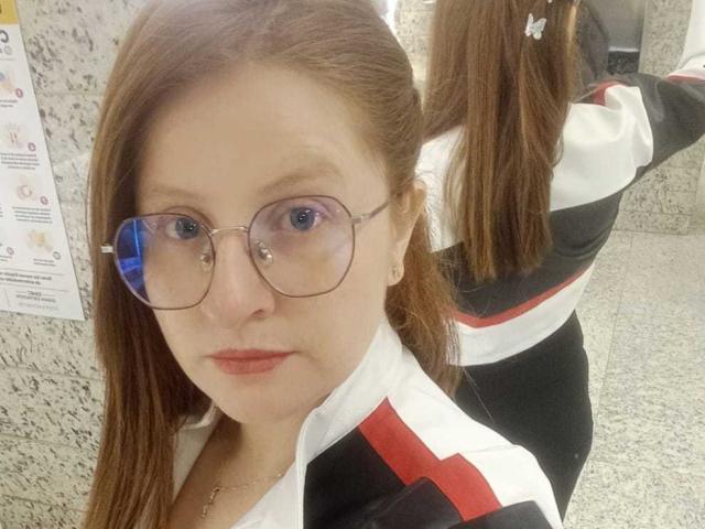 EnzumGinger with red lipstick, glasses, and brown hair in a white, black, and red track suit, showing her ass in the mirror, granite wall in the background.